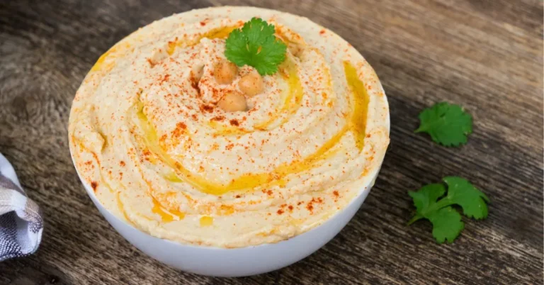 A creamy bowl of hummus garnished with olive oil, paprika, and fresh parsley, served with pita bread and vegetables.